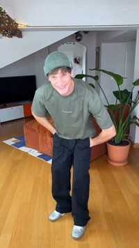 Roheline kootud müts, oliivroheline logoga t-särk, mustad laiad püksid ja hõbedased tossud. Siseala taimedega ja mööbliga.