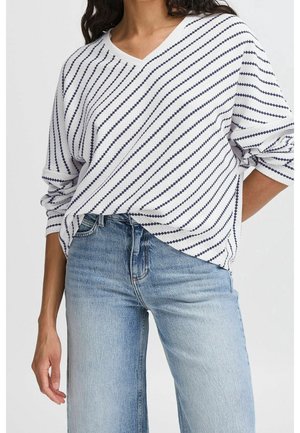 Vrouw in een witte lange mouwen V-hals trui met diagonale marineblauwe strepen, gestoken in lichtblauwe high-waisted jeans met opgerolde mouwen.