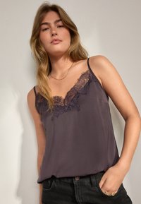 Camisole viola scuro con bordo in pizzo lungo la scollatura e sottili spalline; texture setosa con vestibilità rilassata, abbinata a jeans neri.