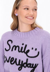 Lila trui met zwarte letters die "Glas elke dag" zegt, met een ribbelachtige hals en zachte breistekstuur. Ronde lavendel oorbellen.