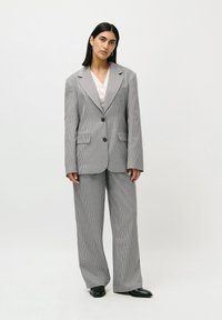 Femeie îmbrăcată într-un blazer supradimensionat în carouri gri și alb și pantaloni largi asortați, purtând pantofi negri, stând pe un fundal alb.