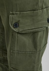 Pantaloni cargo verde oliva realizzati in un tessuto intrecciato e testurizzato. Caratterizzati da una grande tasca laterale con bottone e dettagli di cucitura.