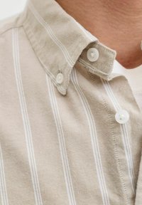 Chemise beige à boutons avec des rayures verticales blanches portée par-dessus un t-shirt blanc à col rond, montrant le col et la partie supérieure de la poitrine.