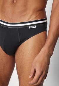 Schwarze Baumwollslips mit einem weißgestreiften Bund, versehen mit einem Etikett. Glattes Material und figurbetontes Design, das die Körperkonturen betont.
