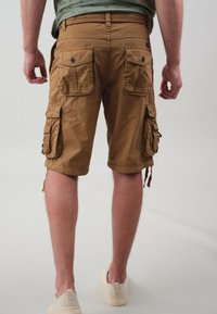 Shorts cargo marron avec plusieurs poches latérales, une coupe décontractée et des cordons de serrage ajustables en bas. Portés avec des chaussures de couleur claire.