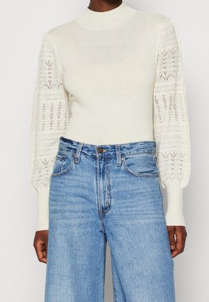 Personne portant un pull blanc avec des manches bouffantes à motifs et un jean bleu taille haute à jambes larges.