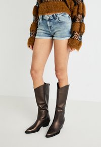 Brūns mākslīgās kažokādas garām piedurknēm tops, gaiši zila denim šorti un mirdzošas, tumšbrūnas līdz ceļiem zābaki ar asu purngali un augstpapēžu zoli.