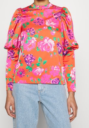 Woman wearing bright orange long-sleeve blouse with pink and purple floral pattern and puffed shoulders, paired with light blue denim jeans.