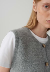Gilet in lana grigia con una morbida e pelosa texture, caratterizzato da un collo a V e due grandi bottoni marroni, indossato sopra una camicia bianca a maniche corte.