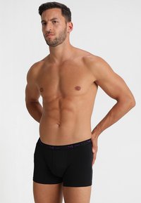 Mustat puuvillaboksereiden alusvaatteet, joissa on purppuranvärinen brändätty vyö, tiukka muotoilu ja sileä pinta. Malli poseeraa kädet lanteilla.