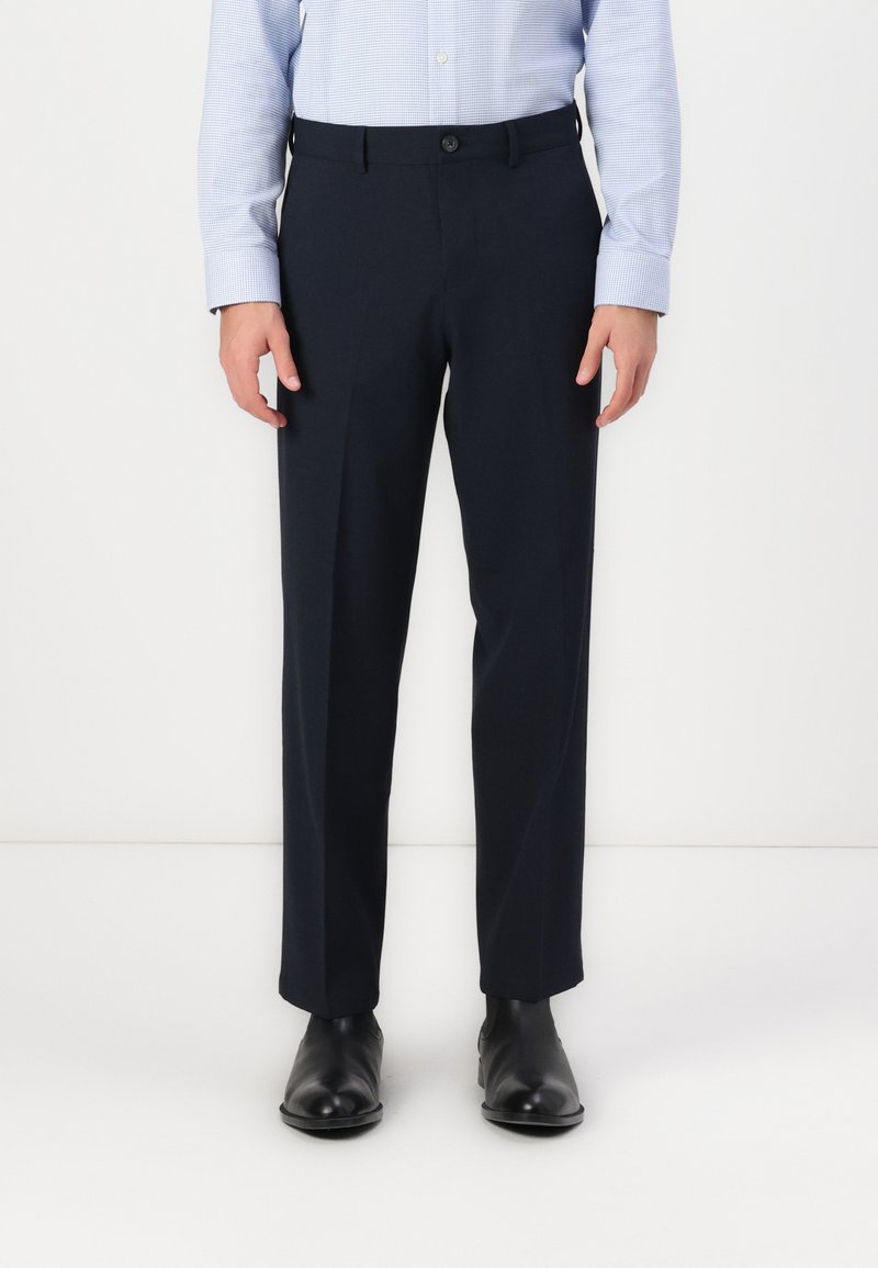Pantalon ajusté bleu marine avec une texture lisse, une coupe droite et une fermeture à bouton, associé à des chaussures noires élégantes.