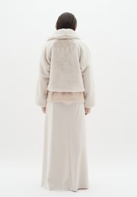 Krämfärgad konstpälsjacka med hög krage, ihop med en lång beige kjol. Mjuk textur och lös passform med sänkt axel.