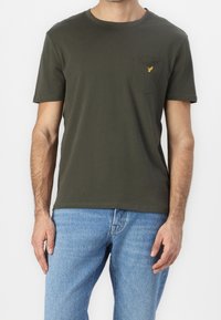 Homme portant un t-shirt à manches courtes vert foncé avec un petit logo jaune sur la poche poitrine et un jean bleu clair.