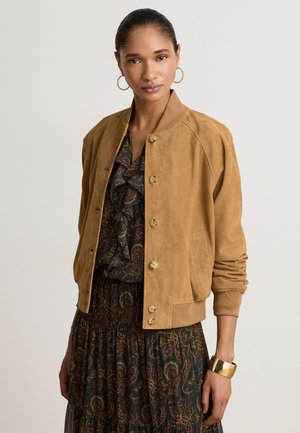 Femme portant une veste bomber marron clair boutonnée sur une robe sombre à motif paisley, des boucles d’oreilles créoles dorées et un large bracelet en or à son poignet.