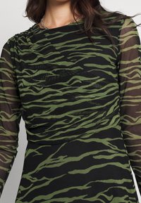 Musta mekko, jossa on pitkät läpikuultavat hihat ja koko pinnan kattava vihreä tiikeriraidoitus. Kangas on sileä ja siinä on roikkuva yksityiskohta kaula-aukossa.