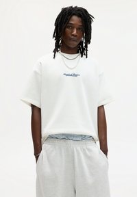 White oversized t-shirt with "Master of Masters" text, paired with light grey sweatpants. Model wears layered necklaces and has dreadlocks.