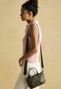 Mała oliwkowozielona torebka z brązowymi skórzanymi elementami, krótkim uchwytem i odpinanym zielonym paskiem na ramię, zestawiona z różowym swetrem na drutach.