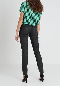 Mujer con cabello rizado que lleva una camisa verde con lunares, pantalones negros ajustados y tacones altos negros, de pie de espaldas.