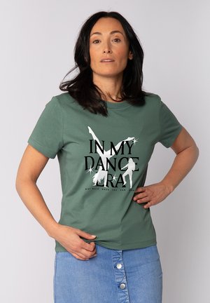 Vrouw met donker haar, gekleed in een groen T-shirt met danssilhouetten en de tekst "IN MY DANCE ERA" en een blauwe, met knopen sluitende spijkerrok.