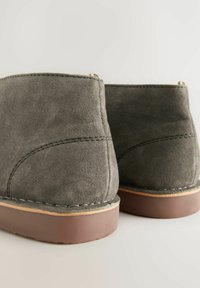 Botas de tobillo de ante gris con una textura suave, suelas de goma marrón en contraste y detalles de costura visibles a lo largo de las costuras.