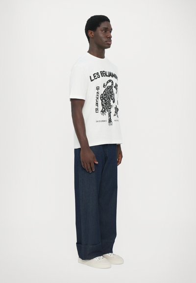 T-shirt blanche avec un motif graphique noir représentant un lion, associée à un jean en denim large bleu foncé et des baskets blanches.