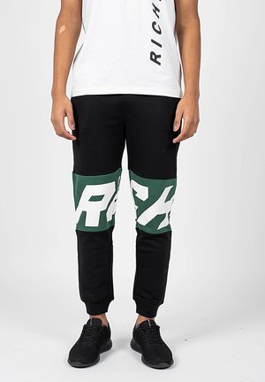 Personne portant un jogging noir avec un motif de lettres vertes et blanches aux genoux, des baskets noires et un t-shirt blanc avec un texte vertical.