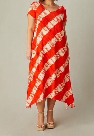 Vrouw draagt een oranje-wit tie-dye korte mouw jurk met asymmetrische zoom en beige plateausandalen op een lichte achtergrond.