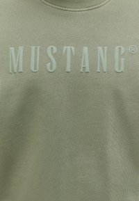 Tissu vert avec le texte en relief "MUSTANG®" vert clair centré sur la zone de la poitrine.