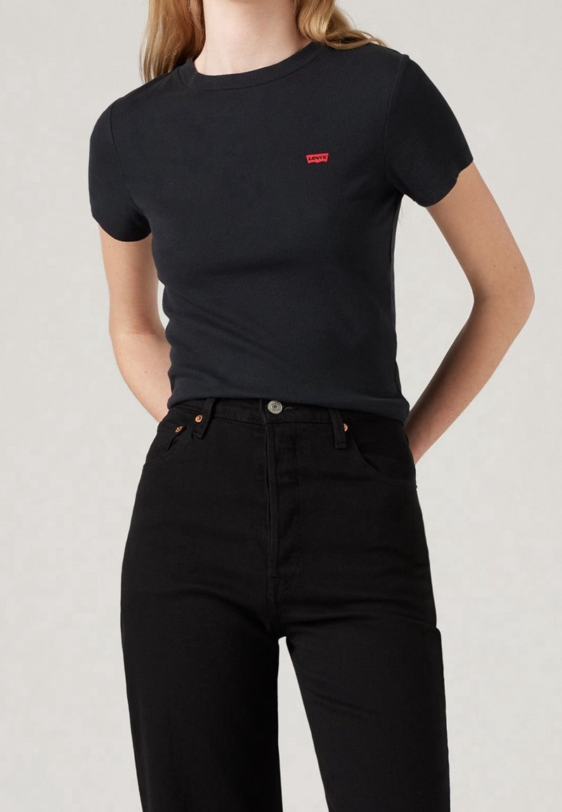 Personne portant un t-shirt noir ajusté à manches courtes avec un petit logo rouge Levi's et un pantalon noir taille haute sur un fond uni.