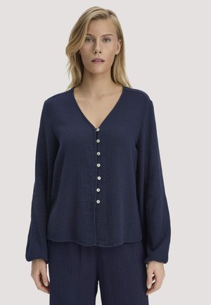 Frau trägt eine locker sitzende, strukturierte dunkelblaue Bluse mit langen Ärmeln, sieben vorderen Knöpfen und passende Hose vor einem einfarbigen hellen Hintergrund.