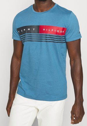 T-shirt en coton bleu avec un col rond, présentant une bande horizontale noire et rouge avec le texte "TOMMY HILFIGER" et des rayures parallèles.