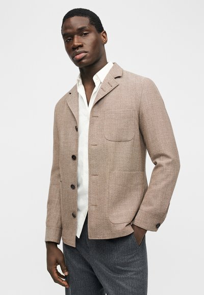 Άνδρας που φοράει μπεζ σακάκι με κουμπιά πάνω από ένα ξεκούμπωτο λευκό πουκάμισο και γκρι παντελόνι, στέκεται με το ένα χέρι στην τσέπη μπροστά σε απλό φόντο.