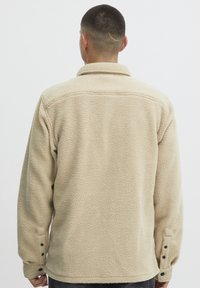 Beige fleecejakke med teksturert overflate, krage og knappede mansjetter, vist fra baksiden. Uformell design som er egnet for lagpålegging.