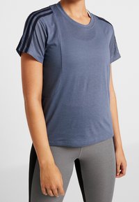 Modrá sportovní tričko s síťovinovými panely na rukávech, kulatým výstřihem a úzkým střihem. V kombinaci se šedými legínami s tmavými bočními akcenty.