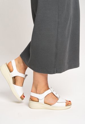 White leather sandals with a T-strap design, open toe, and a cushioned wedge sole. Worn with a gray, ribbed fabric dress.