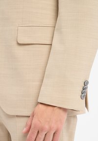 Beżowa marynarka z teksturowanej tkaniny, z wyciętym klapą i pięcioma szarymi guzikami na mankiecie. Po bokach widoczna kieszeń.