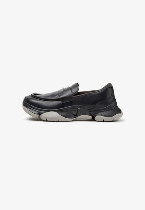 Zapato negro de cuero sin cordones con suela gruesa gris y negra, puntera redondeada y una amplia tira en la parte superior sobre fondo blanco.