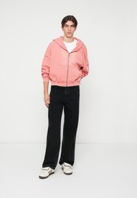 Joven de pie vistiendo una sudadera rosa con cremallera, camiseta blanca, pantalones vaqueros anchos negros y zapatillas blancas con rayas negras.
