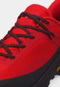 Κοντινό πλάνο κόκκινου αθλητικού παπουτσιού με κορδόνια σε μαύρο και κόκκινο σχέδιο και υφασμάτινο άνω μέρος με υφή, μαύρη σόλα με κίτρινο λογότυπο "Vibram".