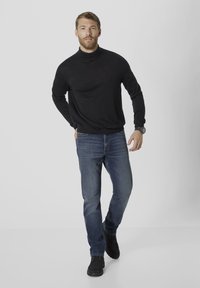Schwarzer Rollkragenspullover, blaue Jeans und dunkles Schuhwerk. Das Modell steht vor einem neutralen Hintergrund und präsentiert ein lässiges Outfit.