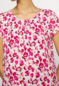 Femme portant un blouse à manches courtes avec un col rond, présentant un motif floral abstrait rose et rouge sur fond blanc.