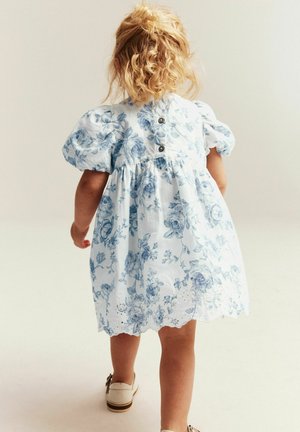 Tout-petit aux cheveux blonds bouclés portant une robe blanche à imprimé floral bleu et des manches bouffantes, marchant de dos sur un fond neutre.