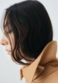 Brown wool coat with a high collar, featuring soft texture and subtle color variations. Dark hair partially frames the face in profile.