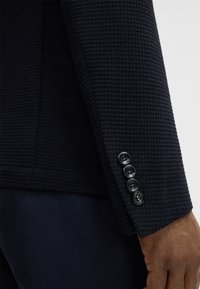 Navyfarbener Blazer-Ärmel mit Struktur, der ein gewebtes Muster und vier schwarze Knöpfe an einem glatten Manschettenaufschlag aufweist.