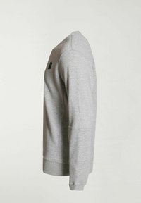 Sudadera gris con cuello redondo, mangas largas y puños de canalé. Presenta un pequeño logo negro en la parte superior izquierda. Textura suave.