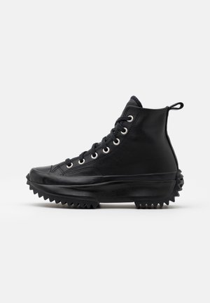 Czarny sneaker na wysokiej cholewce z grubą, postrzępioną podeszwą, sznurowany z przodu z metalowymi oczkami i pętelką z tyłu, widziany z boku.