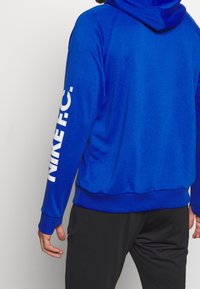 Sweatshirt bleu avec capuche, en tissu texturé. "NINE F.C." imprimé en lettres blanches gras le long de la manche droite.