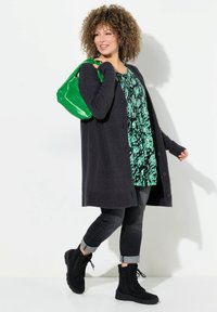 Langer dunkelgrauer Cardigan, florales grün-schwarzes Oberteil, dunkelgraue Jeans, schwarze Schnürstiefel und eine glänzende grüne Handtasche.