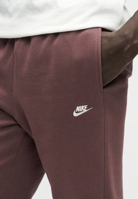 Zbliżenie osoby noszącej bordowe spodnie dresowe Nike z ręką w kieszeni, na tle której widoczny jest biały logo Nike na udzie.