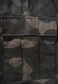 Gros plan d’un tissu camouflage vert foncé et marron avec une poche rectangulaire cousue avec un rabat.